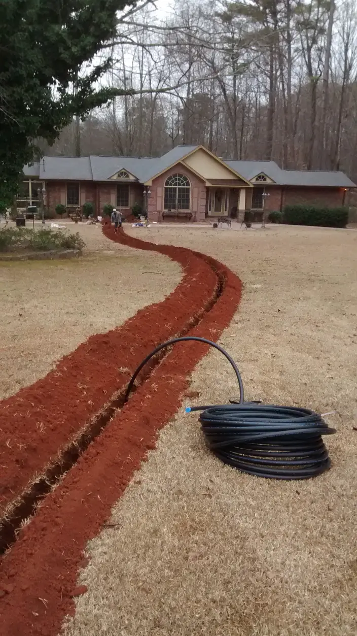A trench cut across a front lawn for a replacement water service line, with the new coiled pipe ready to lay